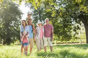 A family of seven, including a child sitting on a man's shoulder, stands in a grassy field with trees in the background.