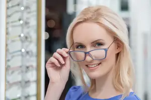 A woman with blonde hair is wearing glasses and is smiling while holding them with both hands