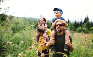 A man carrying a child on his shoulders in a grassy field with another child and woman behind him.