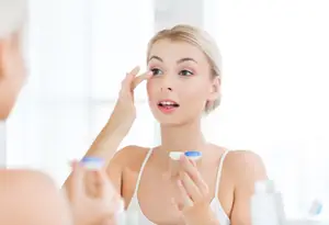 A woman looks at herself in the mirror and holds a pair of contact lenses in her hands.