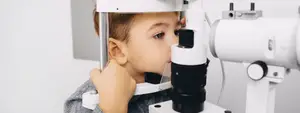 A young boy is looking into an eye examination machine in a clinic room.