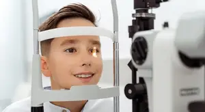 A boy with brown hair is smiling and looking at an ophthalmoscope in a medical setting.