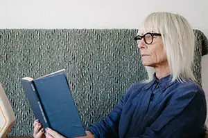 An older woman with white hair wearing glasses is sitting on a couch reading a book.