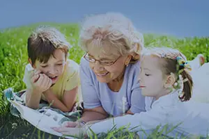 An elderly woman and two children are reading a book in a grassy field under a blue sky.