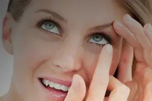 A woman wearing glasses adjusts her contact lens while smiling
