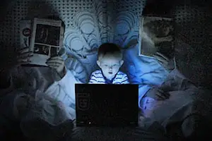 Three people are sitting in a dark room with a laptop on a bed, one person holding a book, another reading a book, and a child looking at the laptop screen