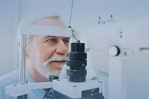 An elderly man with a white beard is wearing glasses and looking through an eye examination machine.