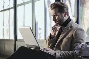 A man sitting on a chair inside a building, using a laptop while looking outside the glass window
