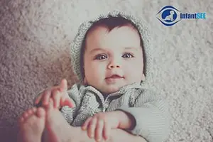 An infant boy in a gray hoodie lies on a carpeted floor, looking directly at the camera.