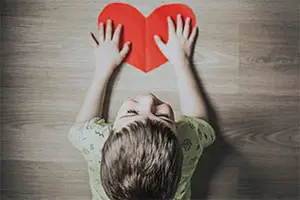 A child holding a red heart shaped paper in both hands, standing on a wooden floor.