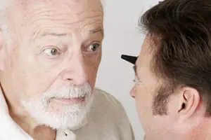 An elderly man with a white beard is being examined by a young man using a black medical tool, possibly a ophthalmoscope, in a room with a white wall.