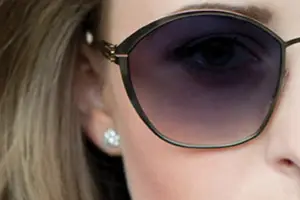 A close-up view of a woman's face, focusing on her eye and sunglasses. The woman has long blonde hair, and she is wearing a pair of black sunglasses with a gold frame and a silver earring. The sunglasses have a gradient lens that transitions from dark to light, and the woman's eye is slightly closed, possibly due to the reflection of the sunglasses.