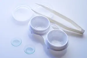 A pair of tweezers, a pair of lenses, and a glass container with a clear liquid are on a white surface.