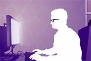 A silhouette of a man sitting in front of a computer monitor with his hand on the keyboard.