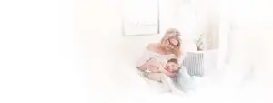 A mother and child are sitting on a bed with pillows and smiling in a white room.