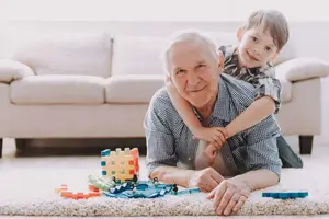 An elderly man lying on the floor with a smiling boy sitting on his chest, surrounded by colorful building blocks, with a white couch in the background.