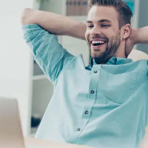 Man smiling and leaning back in a chair with his hands behind his head in front of a laptop