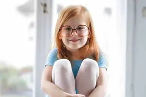 A young girl with red hair is sitting in front of a window, smiling and wearing glasses.