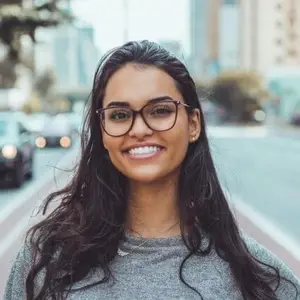 A woman wearing glasses and a gray sweater is smiling while standing on a city street with cars driving behind her.