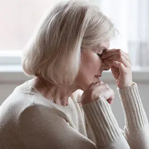An elderly woman with short blonde hair wearing a cream sweater and gold necklace appears to be sneezing or blowing her nose while sitting in front of a window.