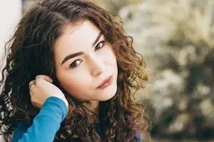 A woman with curly hair wearing a blue long-sleeve shirt is posing for a picture in a garden with plants and trees in the background.