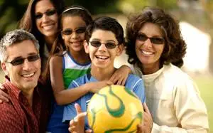 A family of four, including two children, is smiling and posing for a photo in an outdoor setting. They are all wearing sunglasses and the boy is holding a soccer ball.