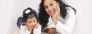 A mother and daughter are lying on a white fur surface, posing for a photo. The mother is wearing a white long-sleeve shirt and has her hands on her cheeks. The daughter has her hands on her face and is wearing a white shirt and jeans.