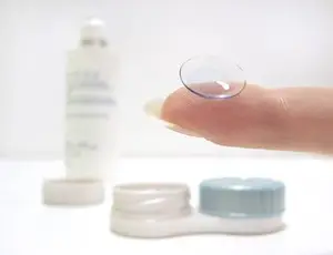 A hand holds a transparent contact lens over a small bottle of eye solution, a container with a blue lid, and another container with a white lid.
