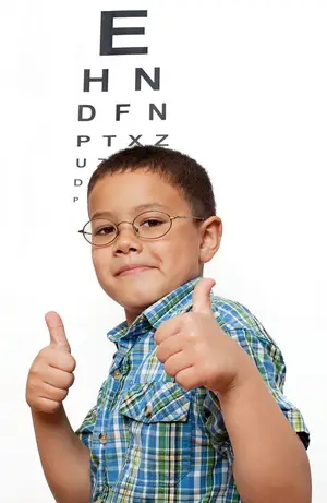 A young boy with glasses gives a thumbs up in front of an eye chart.