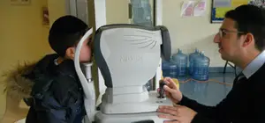 A young boy is undergoing an eye examination using an automatic eye testing machine, with a man operating the machine and the boy standing in front of it.