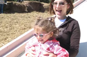 A woman and a young girl are playing on a slide, both wearing sunglasses and smiling, with hay bales and other people in the background.