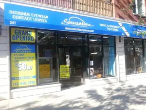 A storefront has a blue awning and yellow signs promoting a grand opening and discounts on designer eyewear and contact lenses. The store offers eye exams for adults and children, and its website is www.sunsetoneeyewear.com.