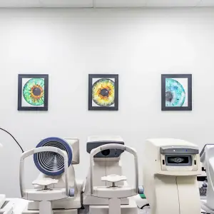 Three framed paintings of eyes hang on a white wall above three medical machines, including a slit lamp and an auto-refractor.