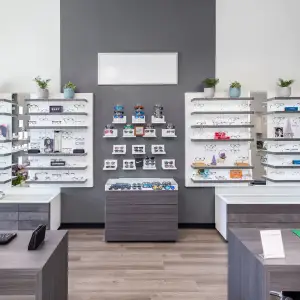 A modern eyeglasses shop interior with wooden floors and gray walls, featuring various eyeglasses on display, gray shelves, and a few potted plants. The shelves have multiple rows of eyeglasses, including some with frames, and the shop has a clean and organized look.
