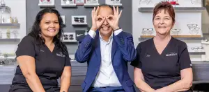 Three people are smiling and posing for a photo in an optical shop, with two women wearing black uniforms and a man in a blue suit, while shelves with items are behind them.