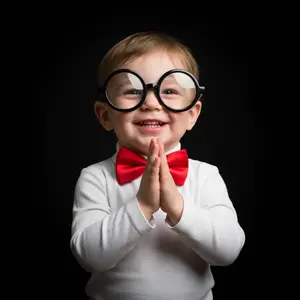 A toddler dressed in a white shirt and red bowtie is wearing oversized glasses and has their hands together in a praying gesture.