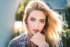 Close-up of a woman with blonde hair, wearing a gray and black patterned cardigan, standing in a garden with a blurred background and a house in the distance.