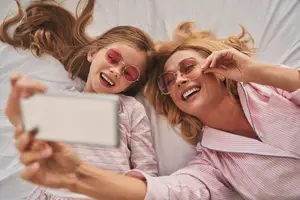 A woman and a girl lying on a bed, smiling and taking a selfie with a phone, both wearing sunglasses.
