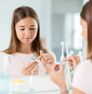 A young woman with brown hair is looking at a toothbrush in front of a mirror, while another person in front of her holds a toothbrush.