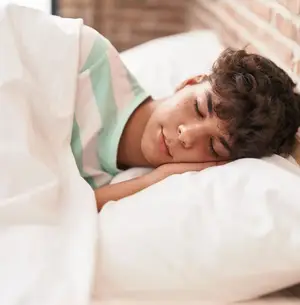 A young boy with curly hair is sleeping on a bed with a pillow and blanket, he is wearing a striped shirt, and his eyes are closed with a slight smile on his face. The boy is lying on his side, and his head is resting on the pillow. The wall behind him is made of bricks, and there is a small window on the left side. The room is dimly lit, and the light is coming from the window.