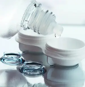 A bottle of contact lens solution is being poured into a lens case with two lenses on a white surface