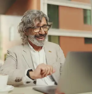 A smiling man sitting at a table with a laptop and a cup of coffee in front of him.