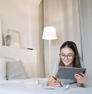 A young girl with glasses is sitting on a bed, holding a tablet and a pen, and smiling while she is doing her homework.