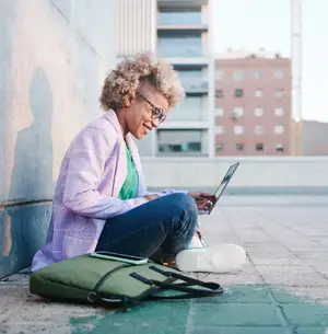 A woman sitting on a sidewalk, smiling and working on a laptop.
