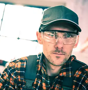 Man wearing a plaid shirt, a cap, and safety glasses with a serious expression