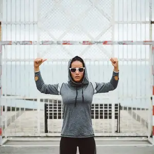 A woman is posing for a photo while wearing a gray hoodie and sunglasses. She is standing in front of a white metal gate. Behind her, there is a bench and a blurry view of the sea and a white fence.