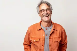 A man with white hair and glasses wearing an orange jacket and gray shirt smiling for a photo