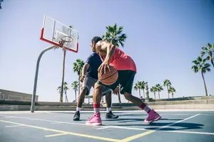 Two men playing basketball on an outdoor court with palm trees in the background.