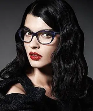 Woman with long black wavy hair wearing a black top and red lipstick, looking forward with glasses on