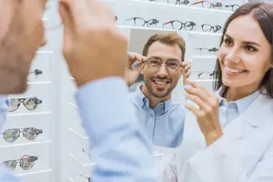 A man trying on glasses at an optometrist's office while a woman looks on and smiles.